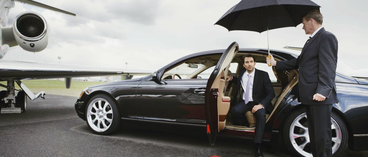 Ein älterer Chauffeur halt seinem Passagier einen Regenschirm hin damit er aus dem Auto aussteigen kann. © Bernd Vogel / Getty Images