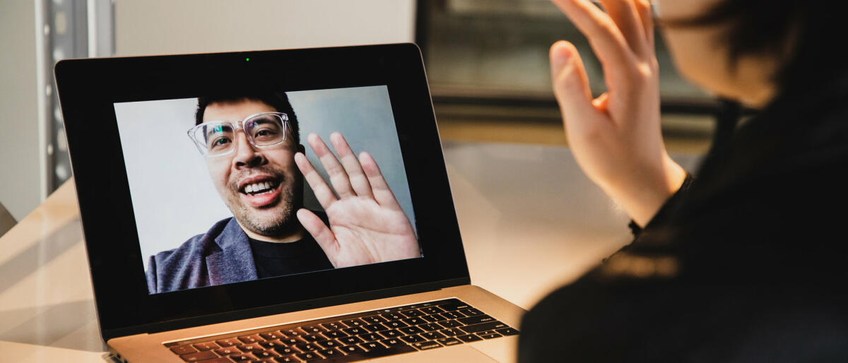 Frau sitzt vor Laptop und winkt Mann in Videokonferenz © Kilito Chan / Getty Images