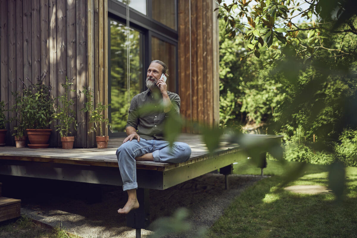 Ein älterer Mann mit grauen Haaren und Zopf sitzt auf einer Terrasse im Grünen und telefoniert lächelnd. © Westend61 / Getty Images