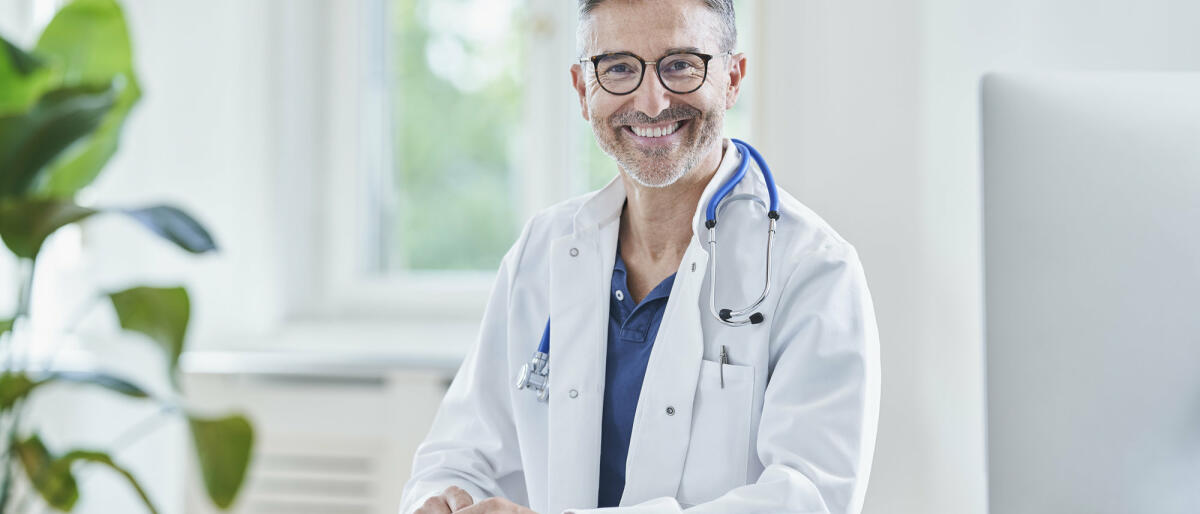 Ein mittelreifer Chefarzt mit weißem Haar sitzt an seinem Schreibtisch und lächelt © Westend61 / Getty Images