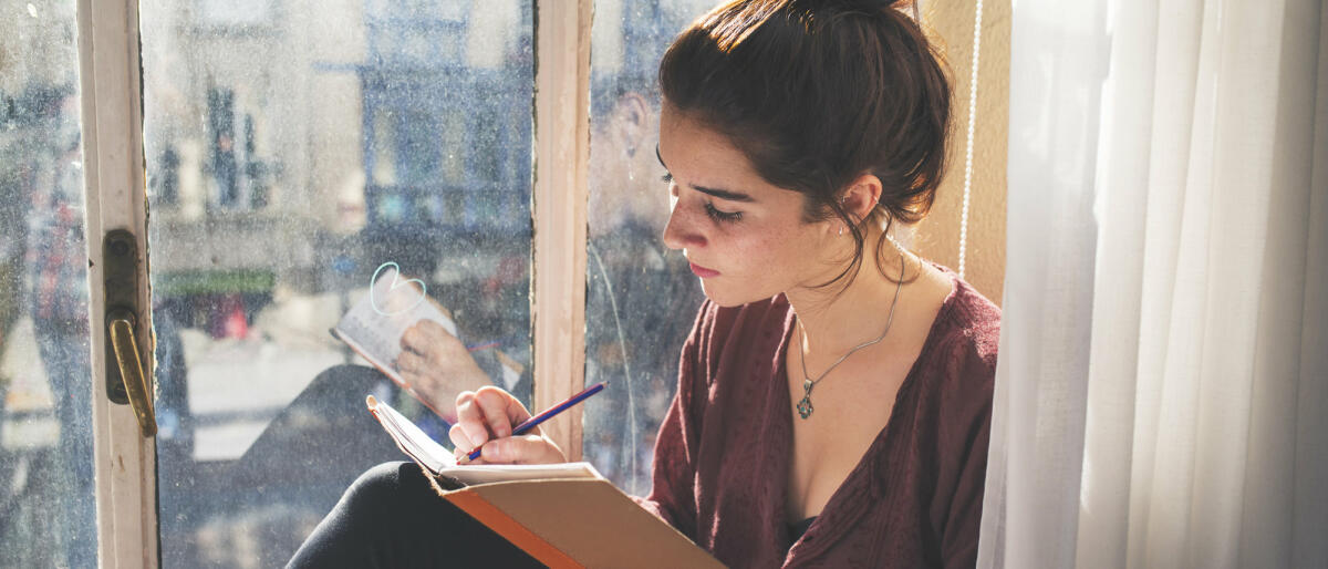 Frau sitzt an Fenster und schreibt in Buch © Leonardo De La Cuesta / Getty Images