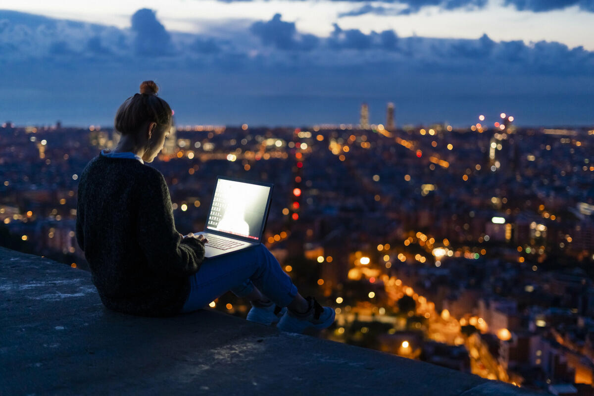 Junge Frau mit Laptop in der Morgendämmerung über der Stadt © Westend61 / Getty Images