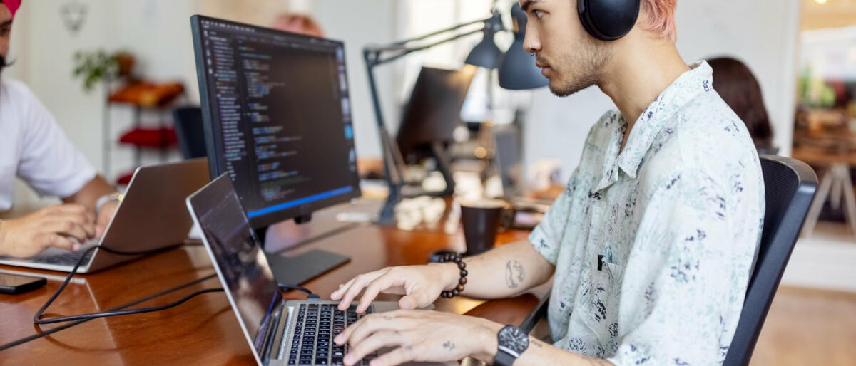 Junger Mann mit Kopfhörern bei der Arbeit am Computer in einem Startup-Büro © Luis Alvarez / Getty Images