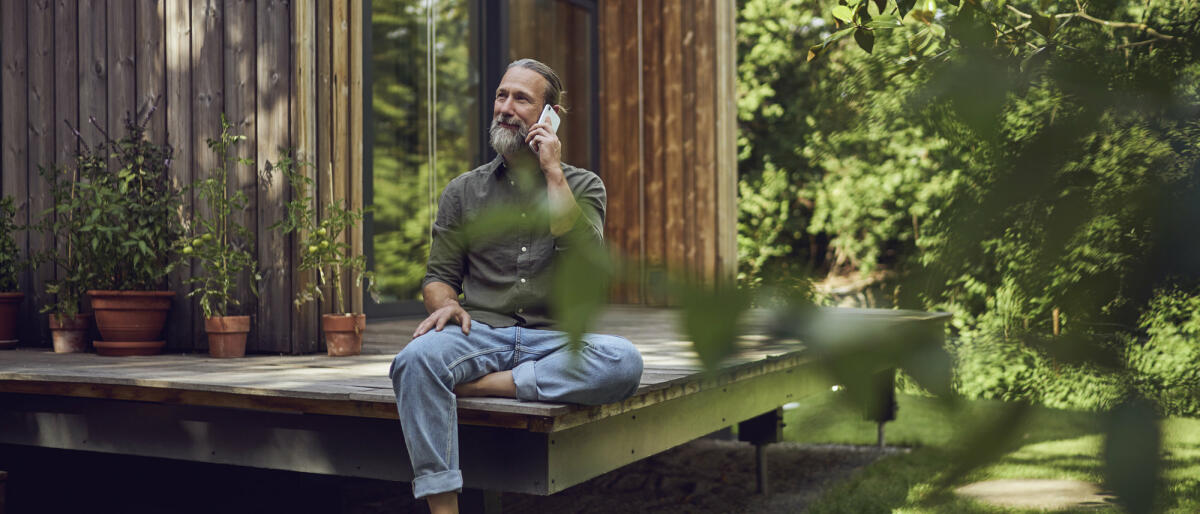 Ein älterer Mann mit grauen Haaren und Zopf sitzt auf einer Terrasse im Grünen und telefoniert lächelnd. © Westend61 / Getty Images