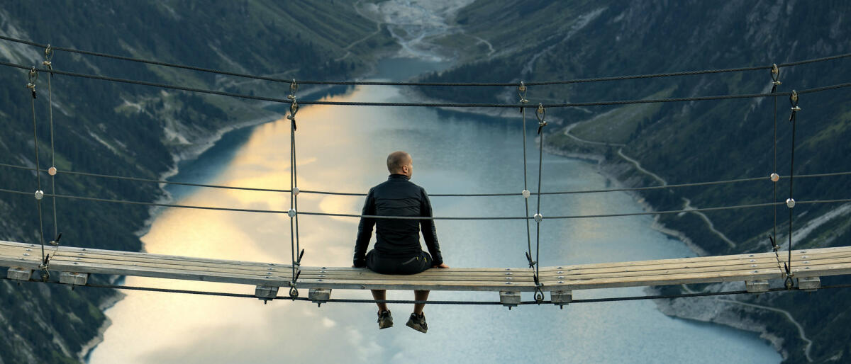 Rückansicht des Mannes, der auf einer Seilbrücke sitzt und die Berglandschaft betrachtet. © ljubaphoto / Getty Images