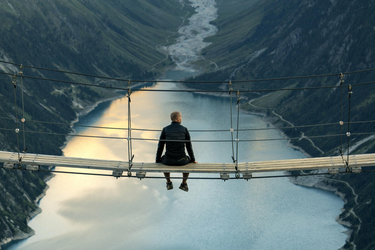 Rückansicht des Mannes, der auf einer Seilbrücke sitzt und die Berglandschaft betrachtet. © ljubaphoto / Getty Images