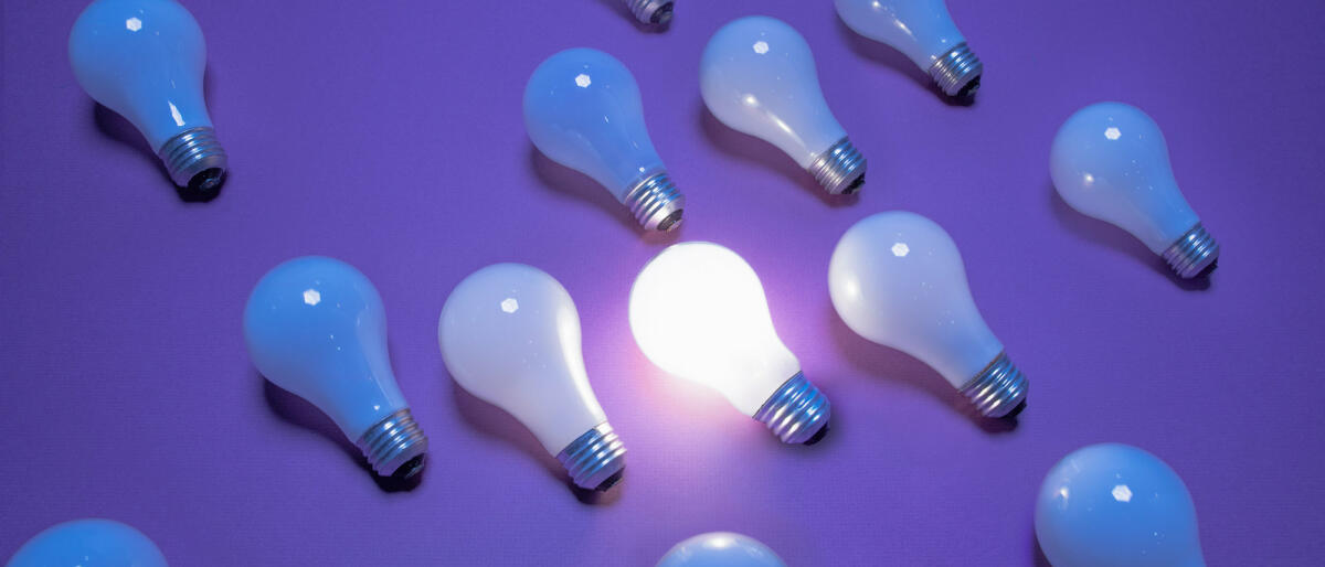 Eine Glühbirne leuchtet zwischen mehreren anderen vor lila Hintergrund © PM Images / Getty Images