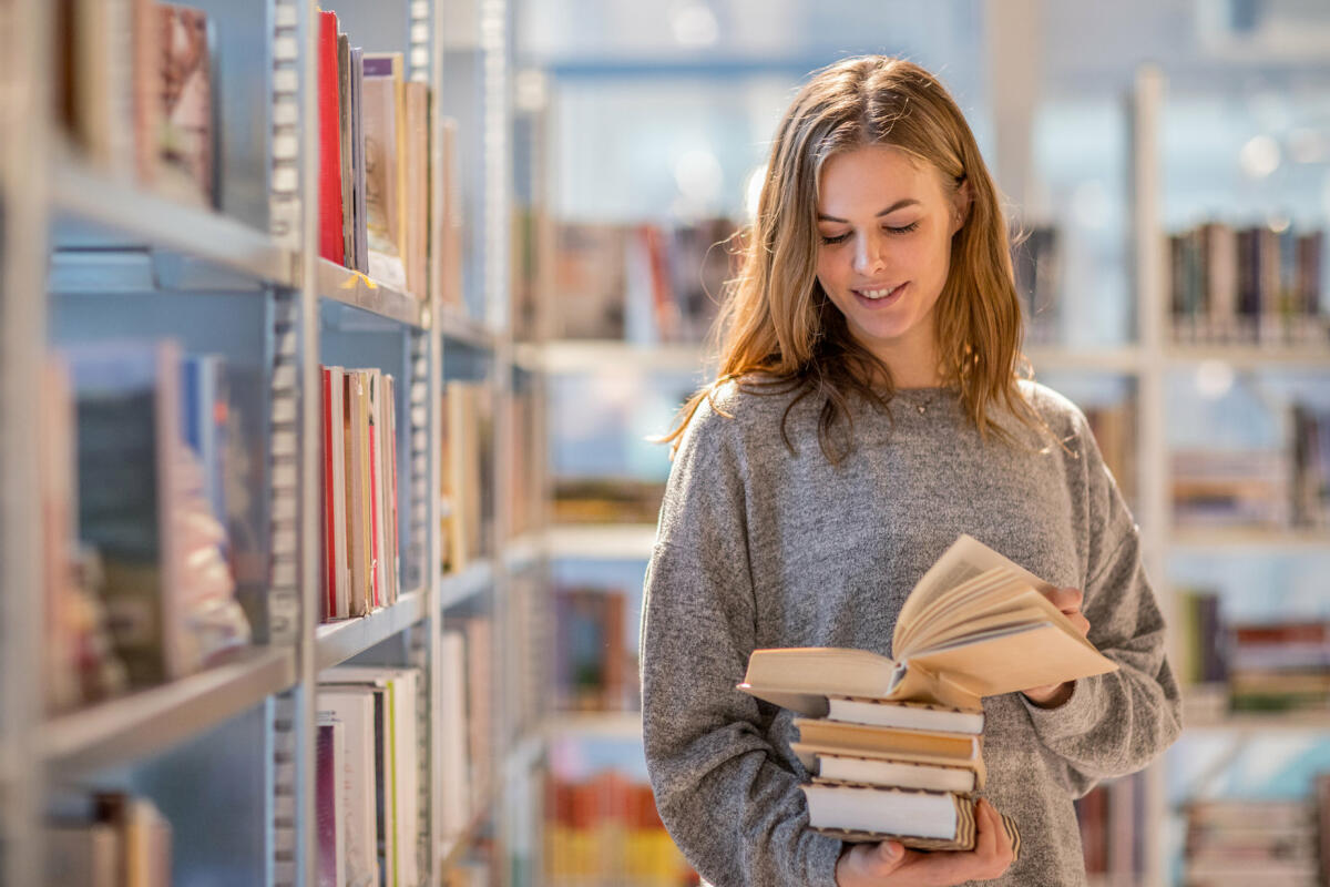 Frau steht in der Bibliothek mit einem Buch in der Hand vor dem Regal © Lorado / Getty Images