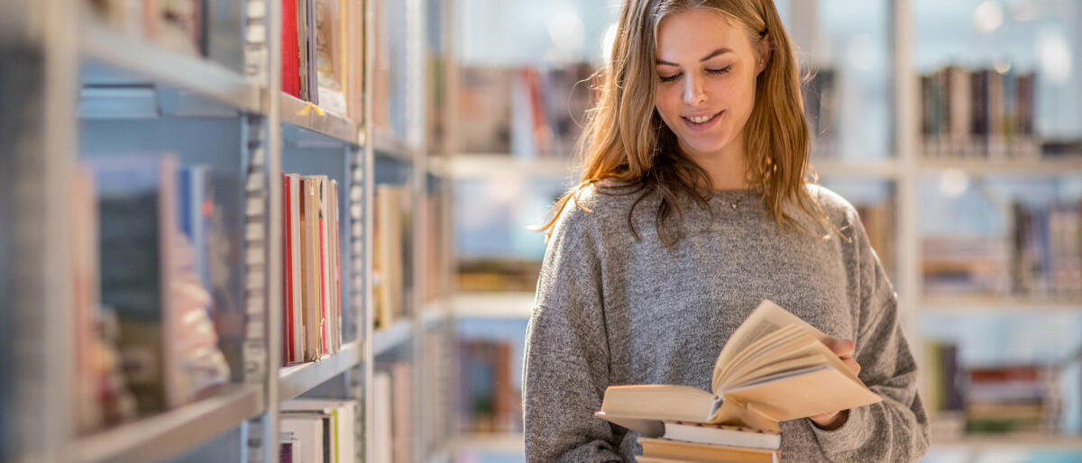 Frau steht in der Bibliothek mit einem Buch in der Hand vor dem Regal © Lorado / Getty Images