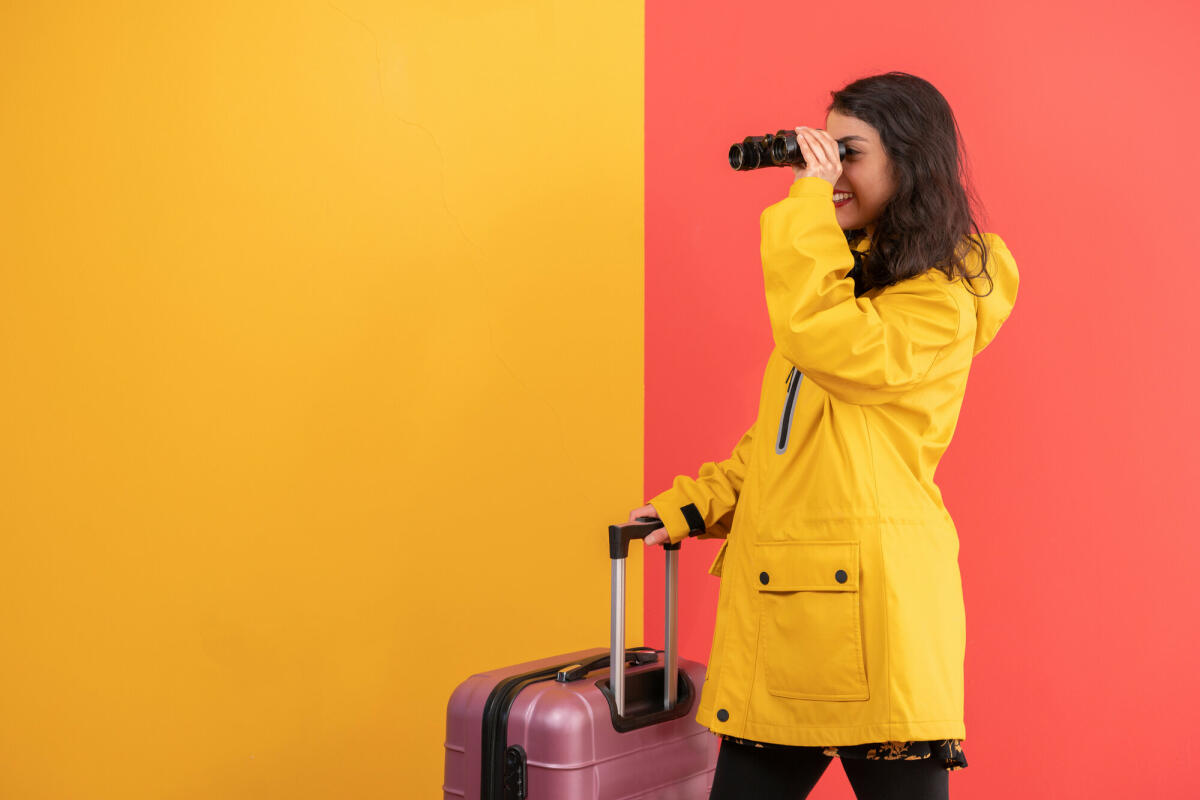 Weibliche Reisende schaut durch Fernglas über farbigen Hintergrund weg. © mgstudyo / Getty Images