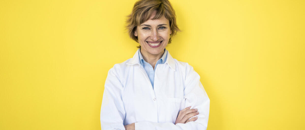 Eine junge Laborantin steht freudig lächelnd vor einem gelben Hintergrund © Westend61 / Getty Imaged