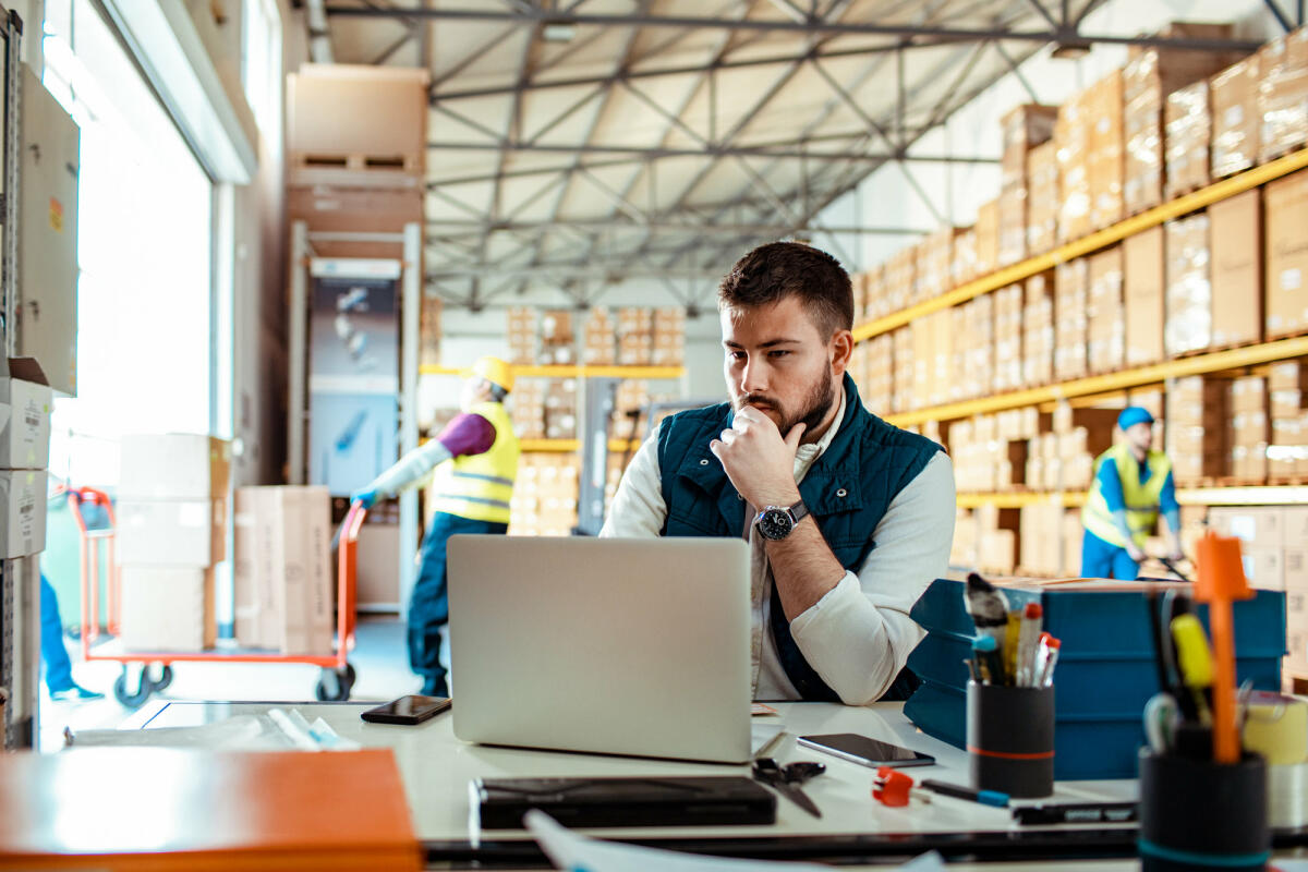 Der Supply Chain Manager sitzt am Laptop in einer Warenversandhalle © Marko Geber / Getty Images
