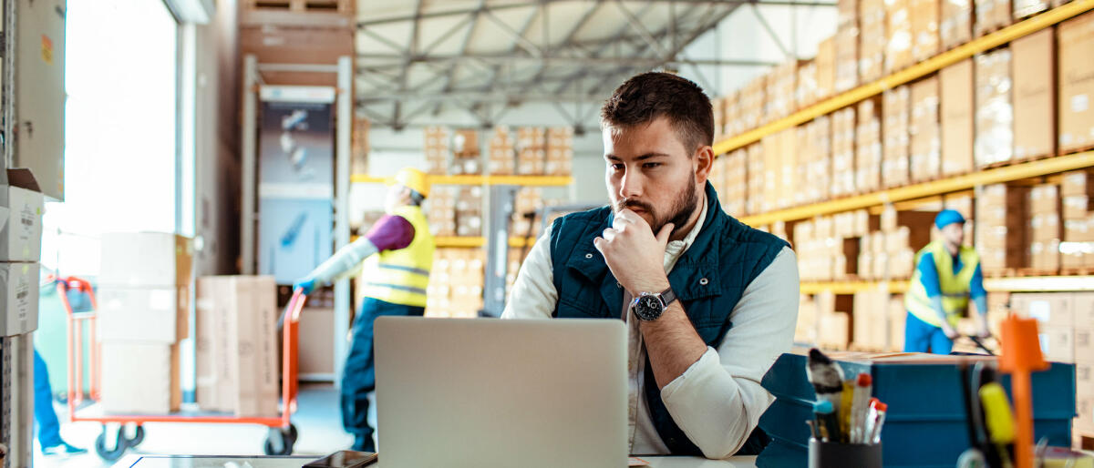 Der Supply Chain Manager sitzt am Laptop in einer Warenversandhalle © Marko Geber / Getty Images
