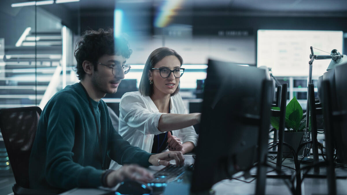 Zwei junge Kollegen, die am Computer arbeiten und sich am Arbeitsplatz unterhalten. Weibliche und männliche Softwareentwickler diskutieren eine Lösung. © gorodenkoff/ Getty Images