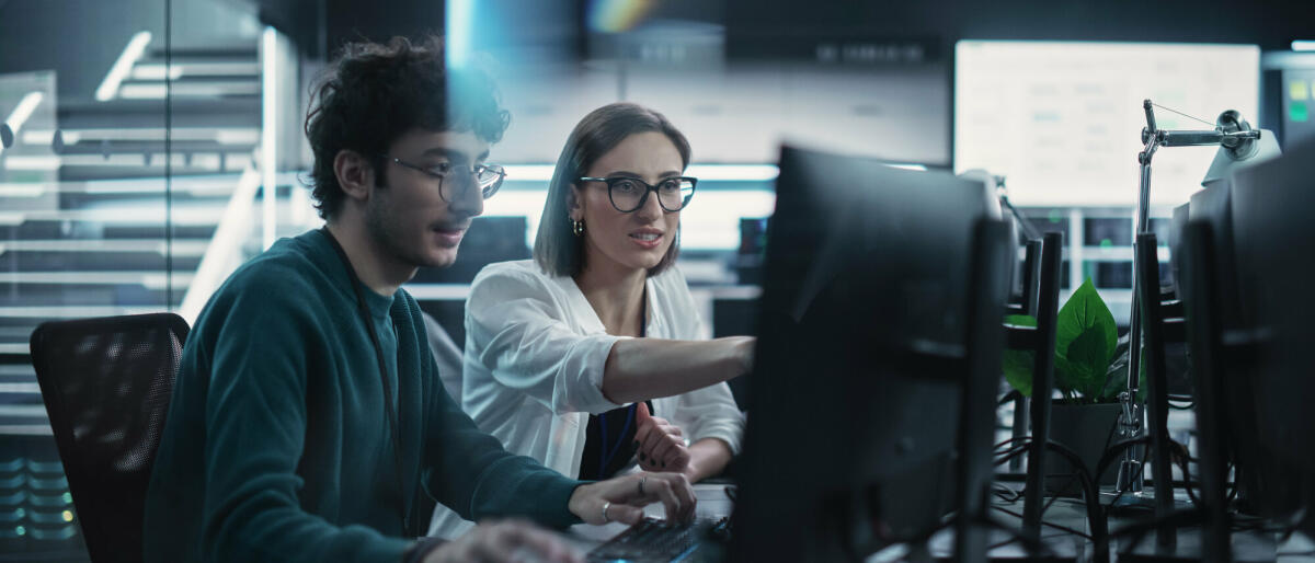 Zwei junge Kollegen, die am Computer arbeiten und sich am Arbeitsplatz unterhalten. Weibliche und männliche Softwareentwickler diskutieren eine Lösung. © gorodenkoff/ Getty Images