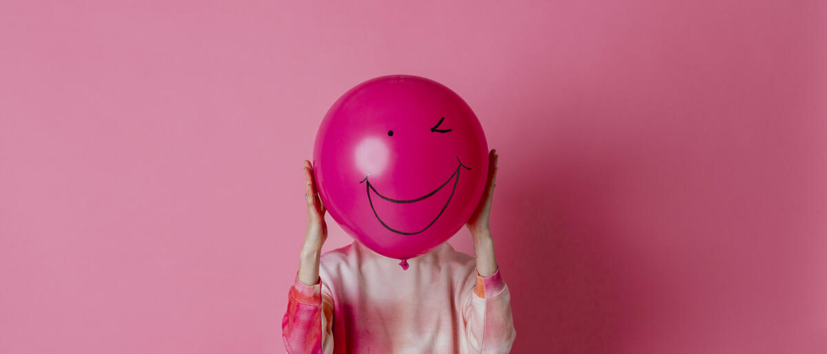 Auf rosa Hintergrund ist ein dreiviertellanges Studioporträt einer jungen Frau in Freizeitkleidung zu sehen. Sie hält einen rosa Ballon mit einem Lächeln. © SolStock / Getty Images