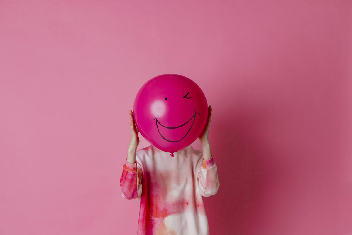 Auf rosa Hintergrund ist ein dreiviertellanges Studioporträt einer jungen Frau in Freizeitkleidung zu sehen. Sie hält einen rosa Ballon mit einem Lächeln. © SolStock / Getty Images