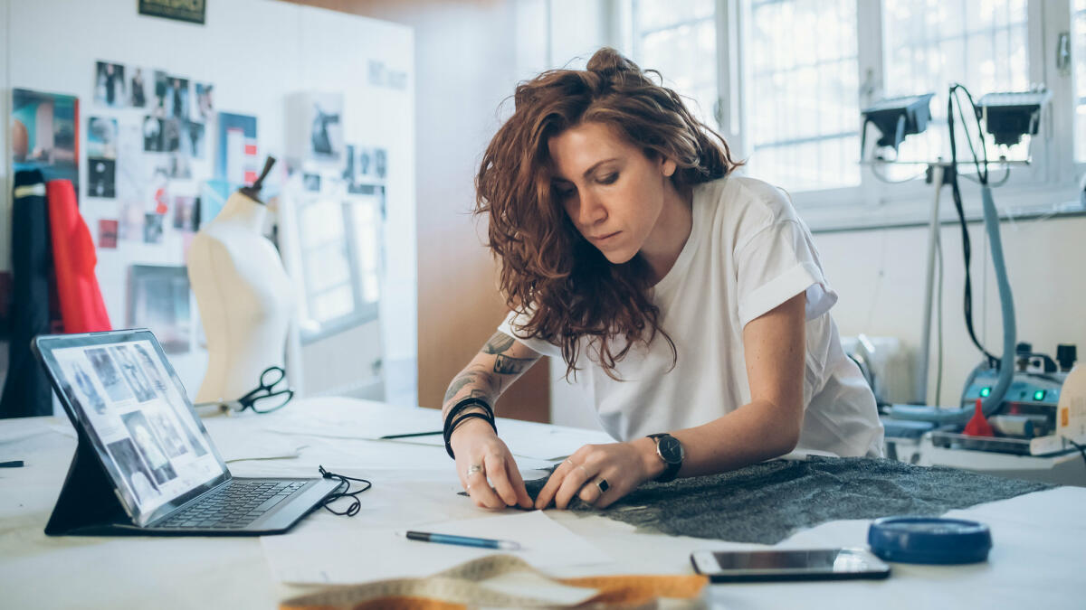 Eine junge tätowierte Textildesignerin beugt sich über ihren Arbeitstisch und arbeitet an einem Stück Stoff © Eugenio Marongiu / Getty Images