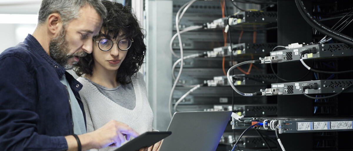 Männlicher Techniker zeigt einer Kollegin ein digitales Tablet. Ingenieur arbeitet mit einem Kollegen am Arbeitsplatz. Sie sind im Serverraum. © Getty Images/ 	Morsa Images
