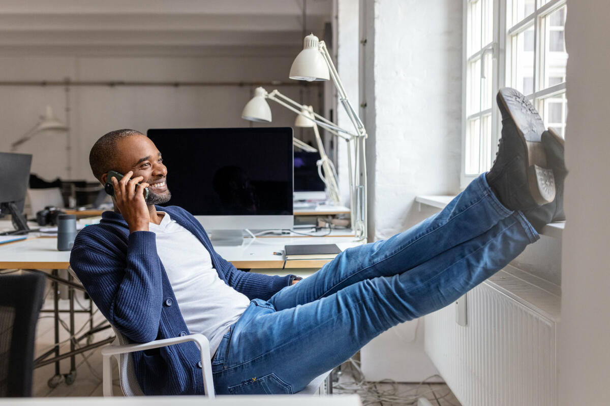 Lächelnder Geschäftsmann, der mit dem Handy telefoniert, während er auf einem Stuhl sitzt und die Füße auf die Fensterbank legt © Luis Alvarez / Getty Images