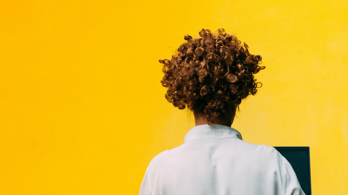 Rückenansicht einer Frau mit kurzen lockigen Haaren am Computer, gelber Hintergrund © Crispin la valiente / Getty Images