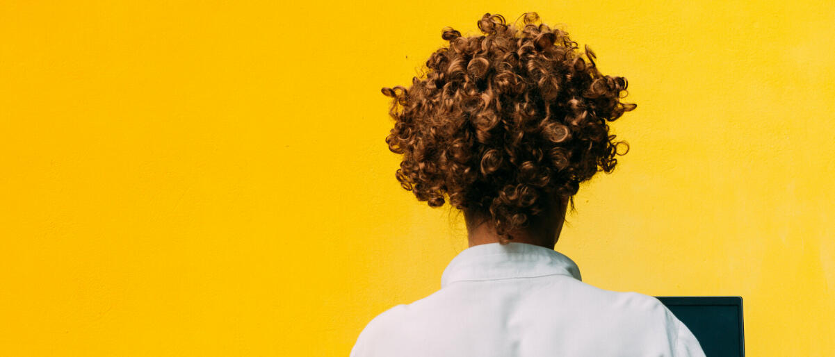 Rückenansicht einer Frau mit kurzen lockigen Haaren am Computer, gelber Hintergrund © Crispin la valiente / Getty Images