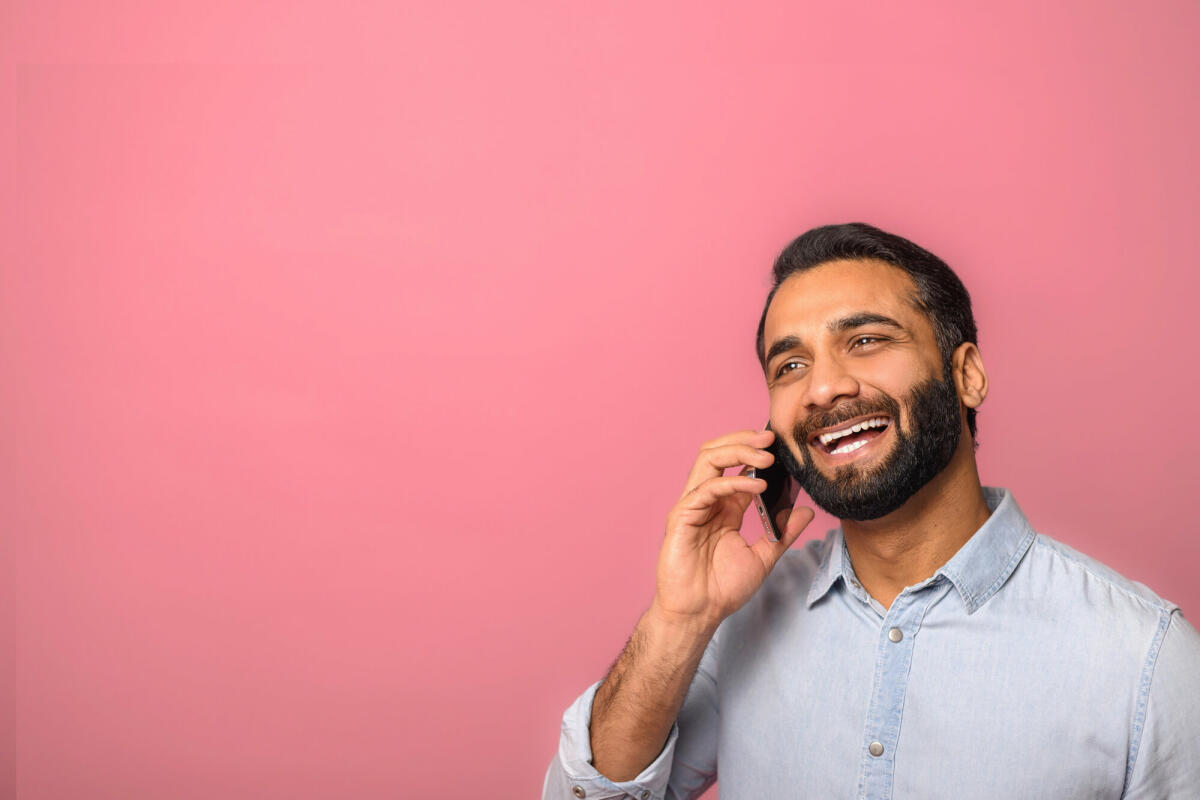 Fröhlicher optimistischer junger indischer bärtiger Mann, der telefoniert, um Pläne zu besprechen, und mit verträumtem Gesichtsausdruck wegschaut. © Vadym Pastukh / Getty Images