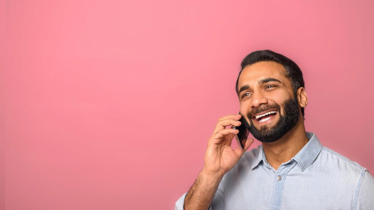 Fröhlicher optimistischer junger indischer bärtiger Mann, der telefoniert, um Pläne zu besprechen, und mit verträumtem Gesichtsausdruck wegschaut. © Vadym Pastukh / Getty Images
