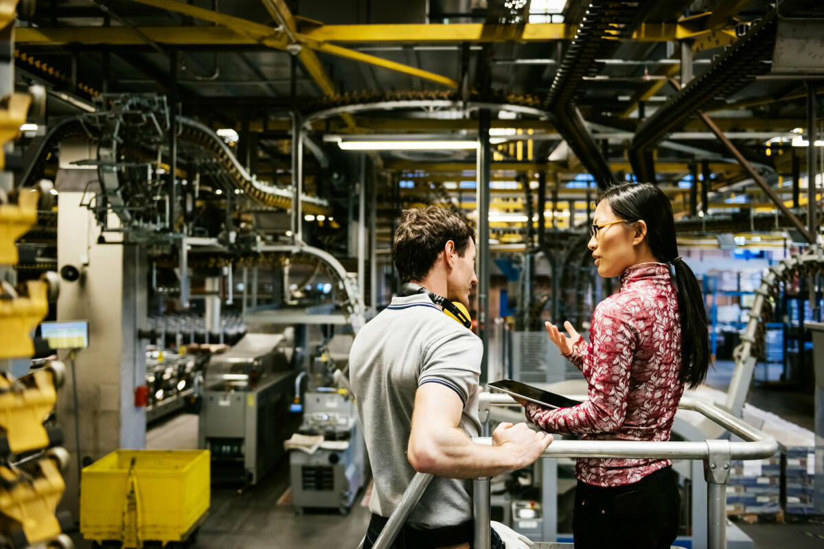 Mechaniker und Manager sprechen in der Nähe einer Maschine in einer großen Druckerei über den neuesten Zeitungsdruck © TommL/ Getty Images