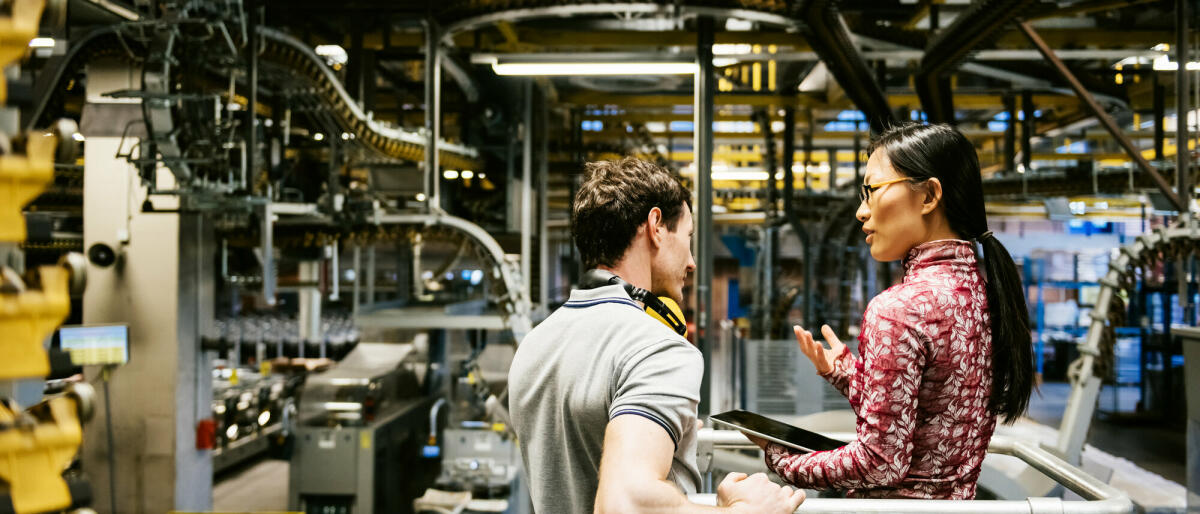 Mechaniker und Manager sprechen in der Nähe einer Maschine in einer großen Druckerei über den neuesten Zeitungsdruck © TommL/ Getty Images