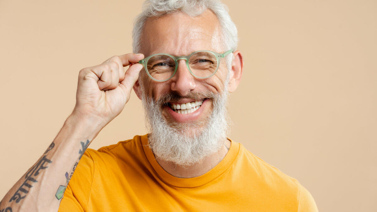 Erfolgreicher reifer Mann, der eine stilvolle Brille und ein gelbes T-Shirt trägt, isoliert auf beigefarbenem Hintergrund © Mariia Vitkovska / Getty Images