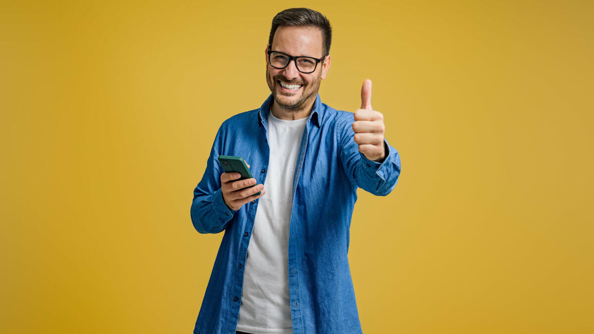 Porträt eines lächelnden Unternehmers, der Daumen nach oben und Nachrichten auf dem Smartphone auf gelbem Hintergrund zeigt © Moon Safari / Getty Images