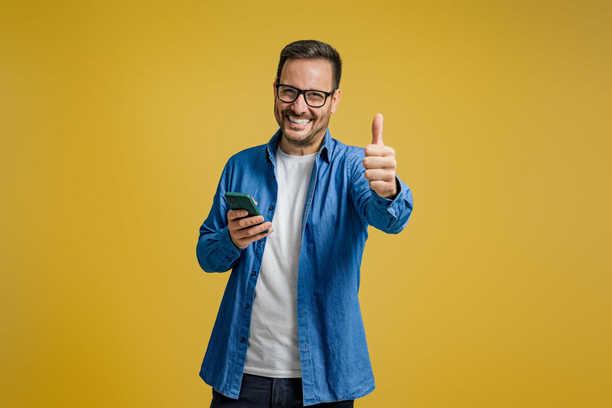 Porträt eines lächelnden Unternehmers, der Daumen nach oben und Nachrichten auf dem Smartphone auf gelbem Hintergrund zeigt © Moon Safari / Getty Images