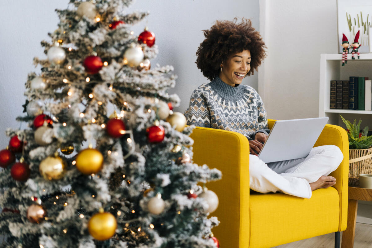 Frau mit Laptop sitzt auf Sessel neben Weihnachtsbaum © Westend61 / Getty Images