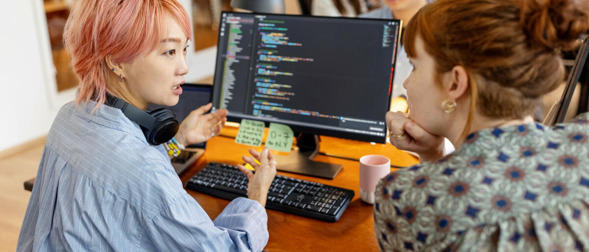 Weibliche Softwareentwicklerinnen sitzen am Arbeitsplatz am Computer und diskutieren kreativ über das neue Programmierprogramm im Büro. © Luis Alvarez/ Getty Images