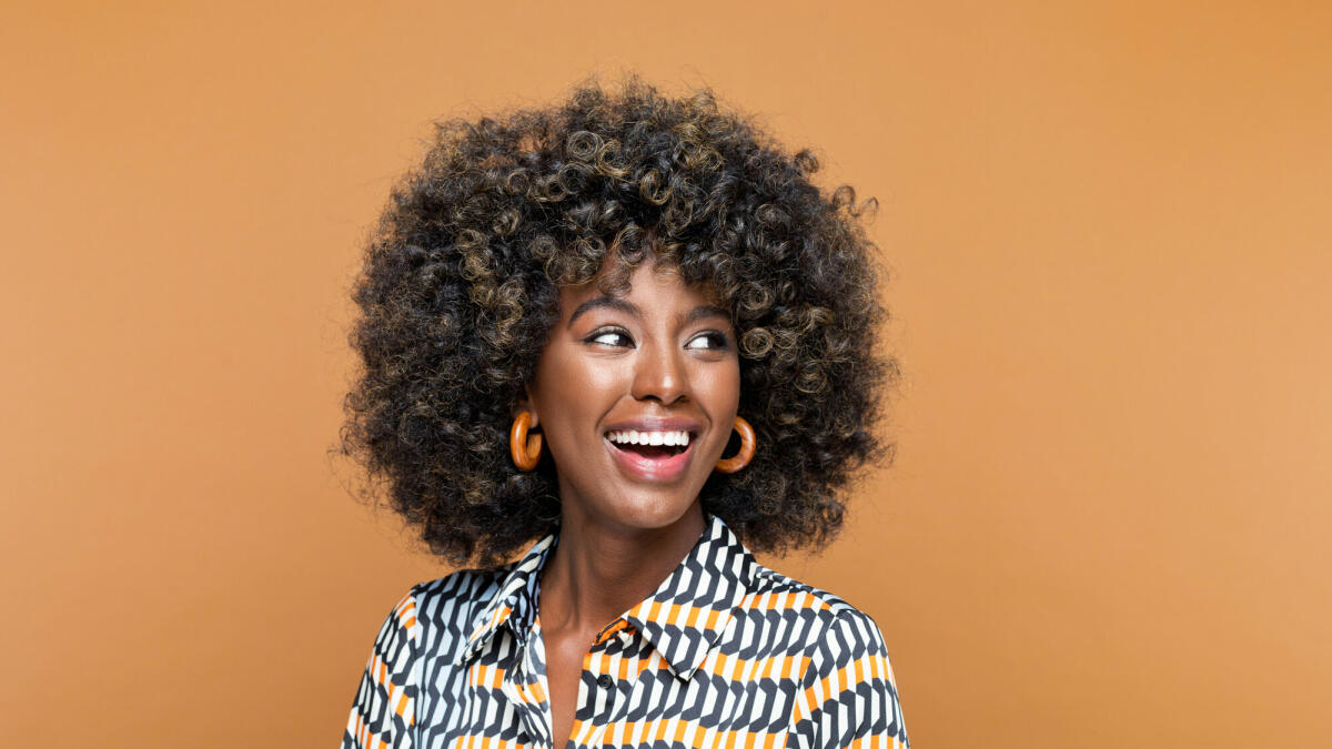 Erstaunte junge Frau mit Afrofrisur, die ein bedrucktes Kleid und hölzerne Ohrringe trägt, wegschaut und lacht. Studioaufnahme auf braunem Hintergrund © izusek / Getty Images