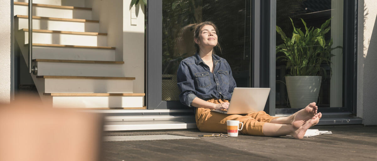 Junge Frau mit Laptop auf dem Schoß sitzt mit geschlossenen Augen in der Sonne  © Westend61 / Getty Images