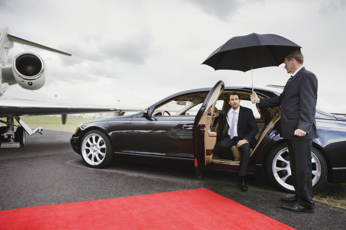Ein älterer Chauffeur halt seinem Passagier einen Regenschirm hin damit er aus dem Auto aussteigen kann. © Bernd Vogel / Getty Images