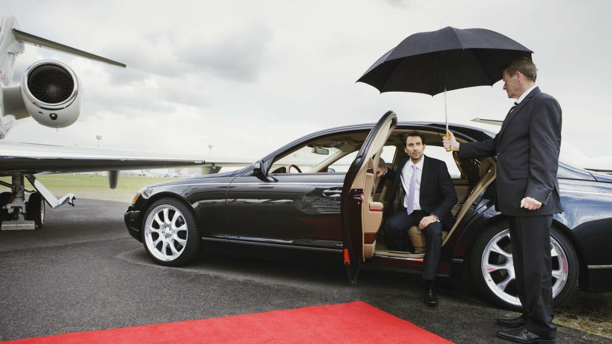 Ein älterer Chauffeur halt seinem Passagier einen Regenschirm hin damit er aus dem Auto aussteigen kann. © Bernd Vogel / Getty Images