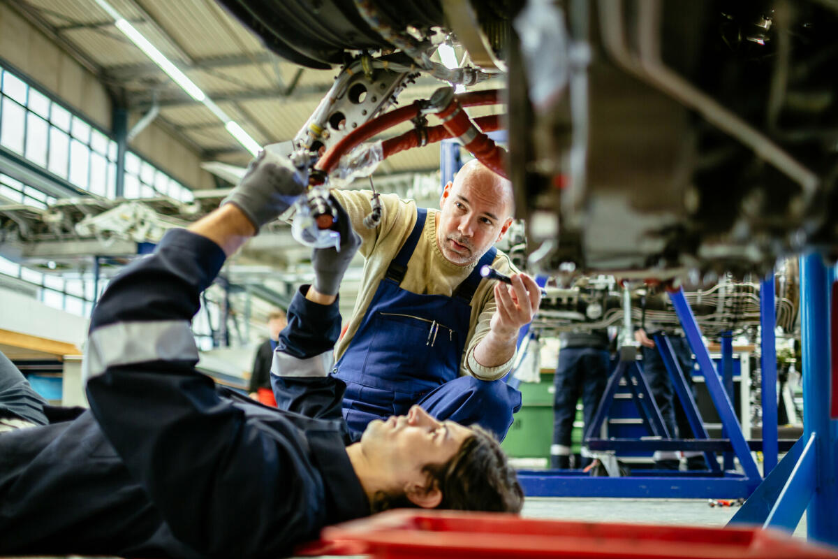 Zwei männliche Flugzeugmechaniker arbeiten an einem Düsentriebwerk in einem Flugzeughangar. Einer von ihnen liegt unter dem Triebwerk, während der andere es mit © Hinterhaus Productions/ Getty Images