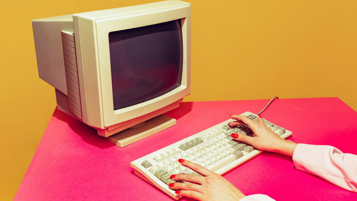 Buntes Bild von Vintage-Computermonitor und Tastatur auf leuchtend rosa Tischdecke über gelbem Hintergrund © master1305 / gettyimages