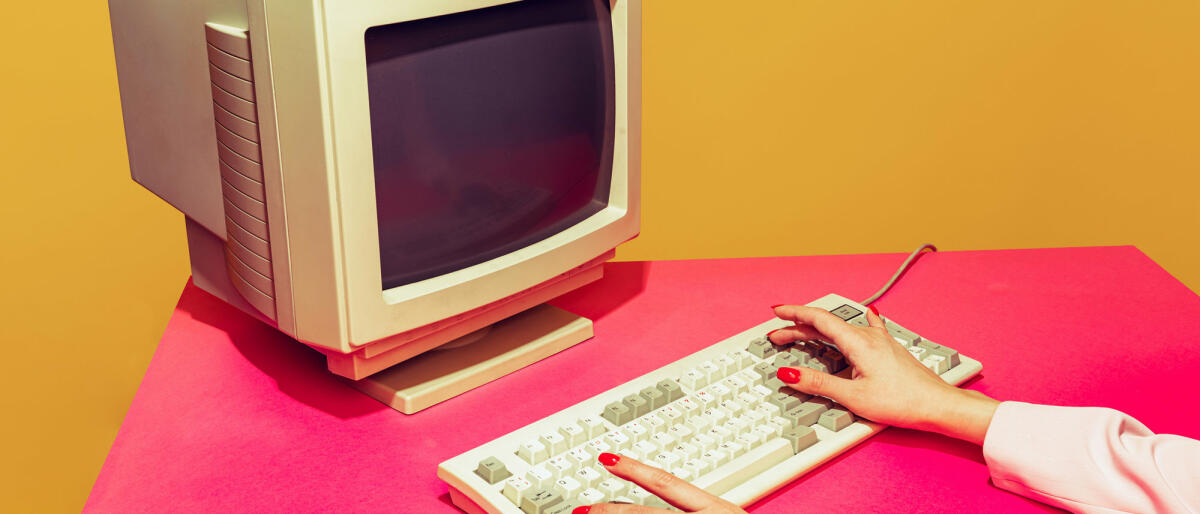 Buntes Bild von Vintage-Computermonitor und Tastatur auf leuchtend rosa Tischdecke über gelbem Hintergrund © master1305 / gettyimages