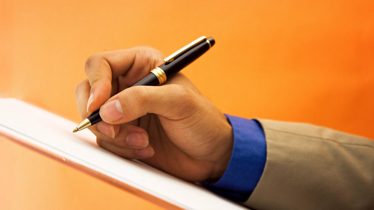 Eine Hand hält einen Kugelschreiber über ein Blatt Papier und setzt zum schreiben an. Der Hintergrund ist einfarbig Orange.  © Nico_Blue / Getty Images
