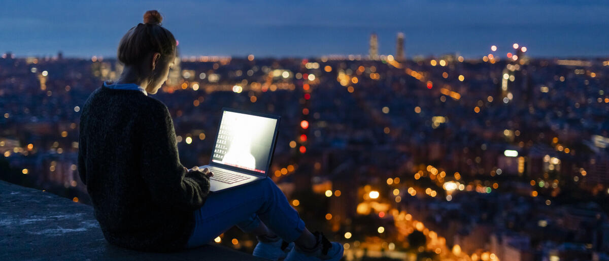 Junge Frau mit Laptop in der Morgendämmerung über der Stadt © Westend61 / Getty Images
