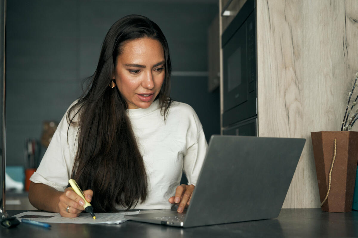 Frau sitzt am Schreibtisch mit Laptop und markiert etwas auf einem Zettel © Sergey Mironov / Getty Images