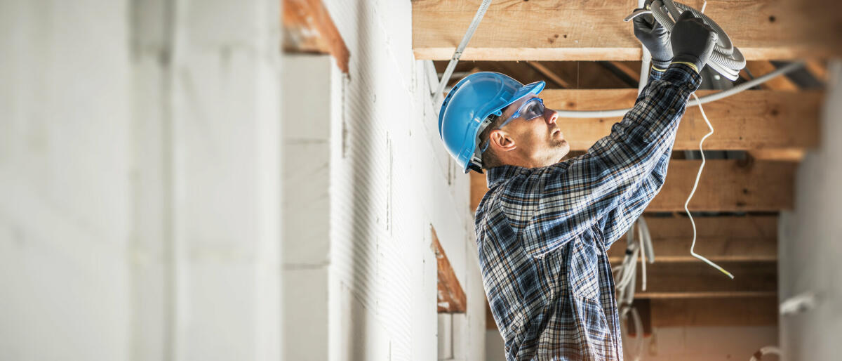 Professioneller kaukasischer Elektriker, der an der Installation der elektrischen Leitungen in der Decke eines neuen Wohnhauses arbeitet. © welcomia/ Getty Images