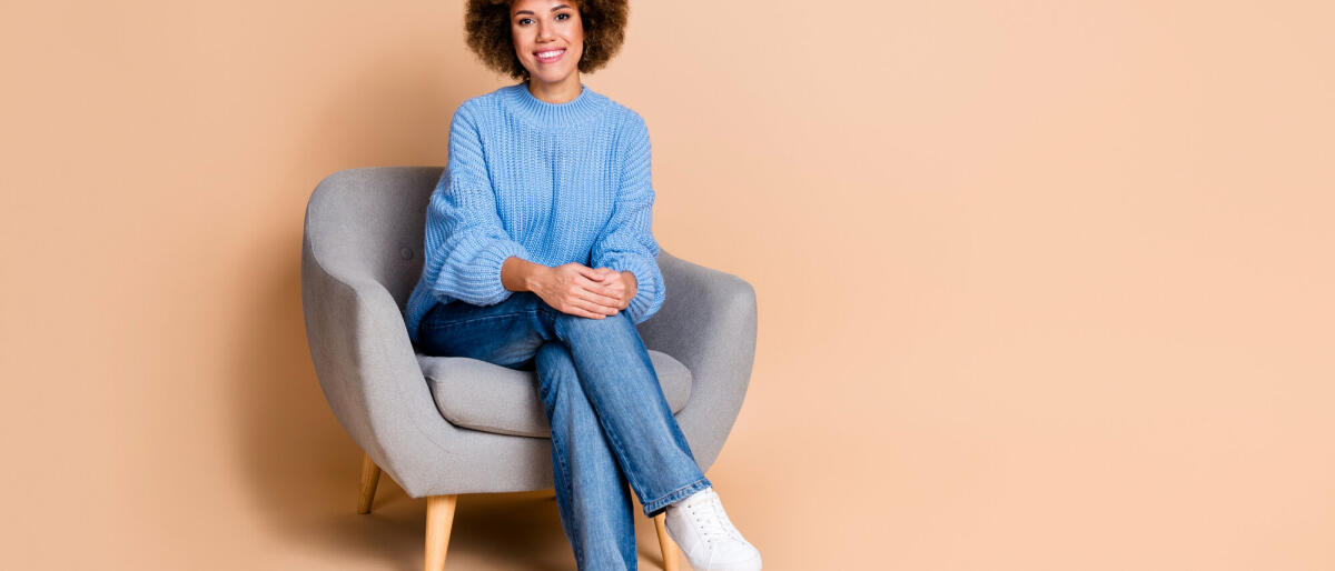 Foto von positivem Mädchen gekleidet strick Pullover Jeans sitzen auf Sessel in der Nähe von Rabatt leerer Raum isoliert auf pastellfarbenem Hintergrund © Deagreez / Getty Images