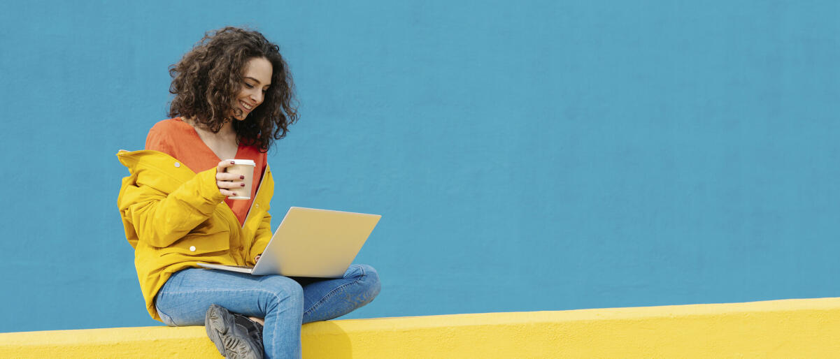 Glückliche junge Frau mit Kaffee zum Mitnehmen, die an einer gelben Wand sitzt und einen Laptop benutzt © Westend61 / Getty Images