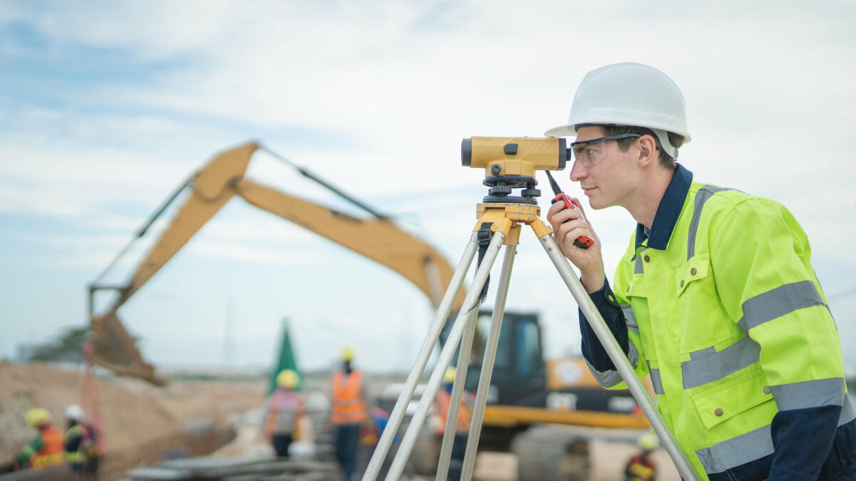 Männlicher Ingenieur bei der Arbeit mit Vermessungsgeräten auf der Baustelle und Arbeiter im Hintergrund © Vithun Khamsong / Getty Images