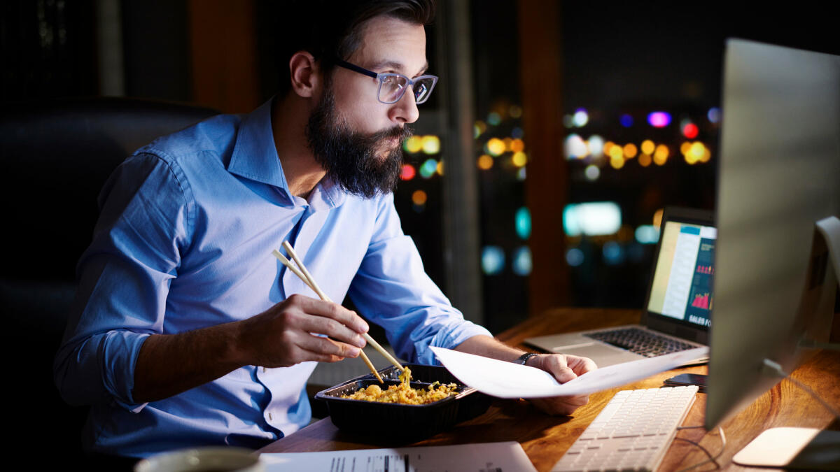 Ein Mann isst nachts vor seinem Computer und macht Überstunden © Gpointstudio / Getty Images